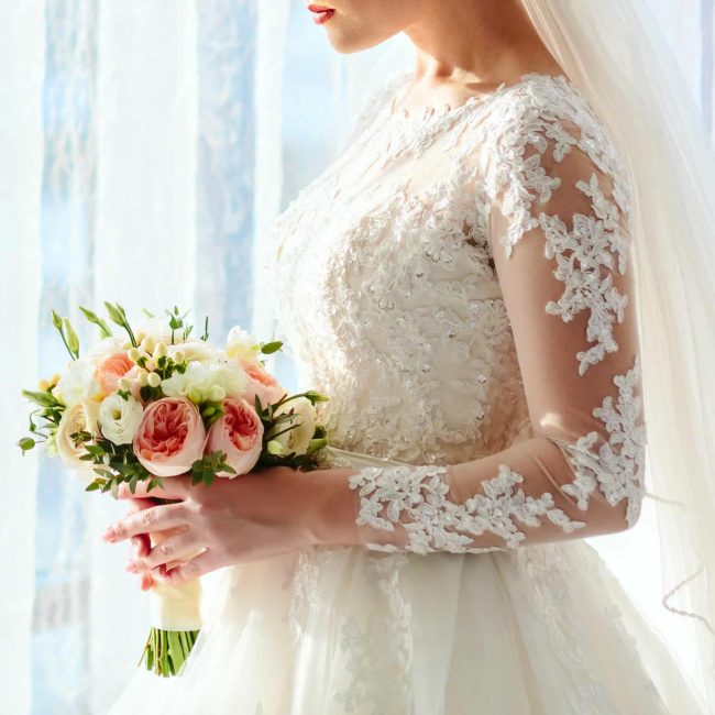 A beautiful bride is holding a wedding bouquet with white roses and peach peonies on a bright window background. Close-up, indoor